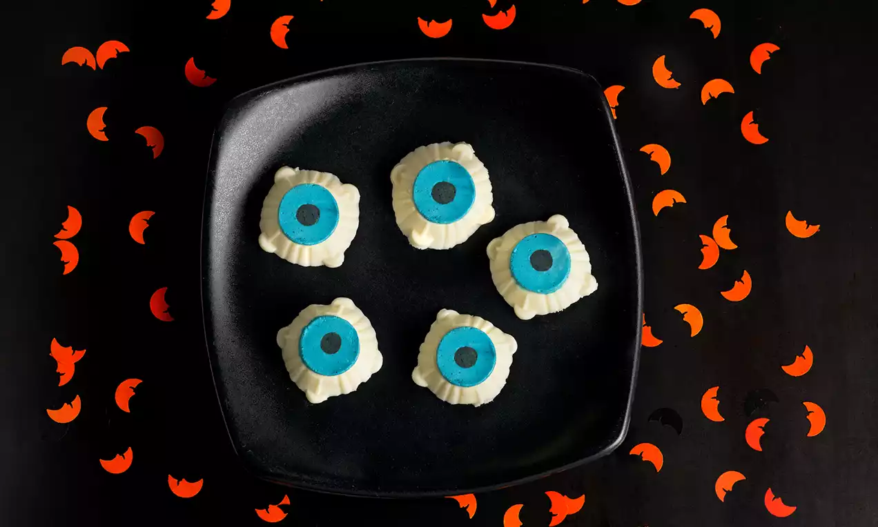 Ojos de gelatina con iris azul y pupila negra sobre esferas blancas de textura suave, dispuestos en un plato negro sobre fondo decorado con murciélagos