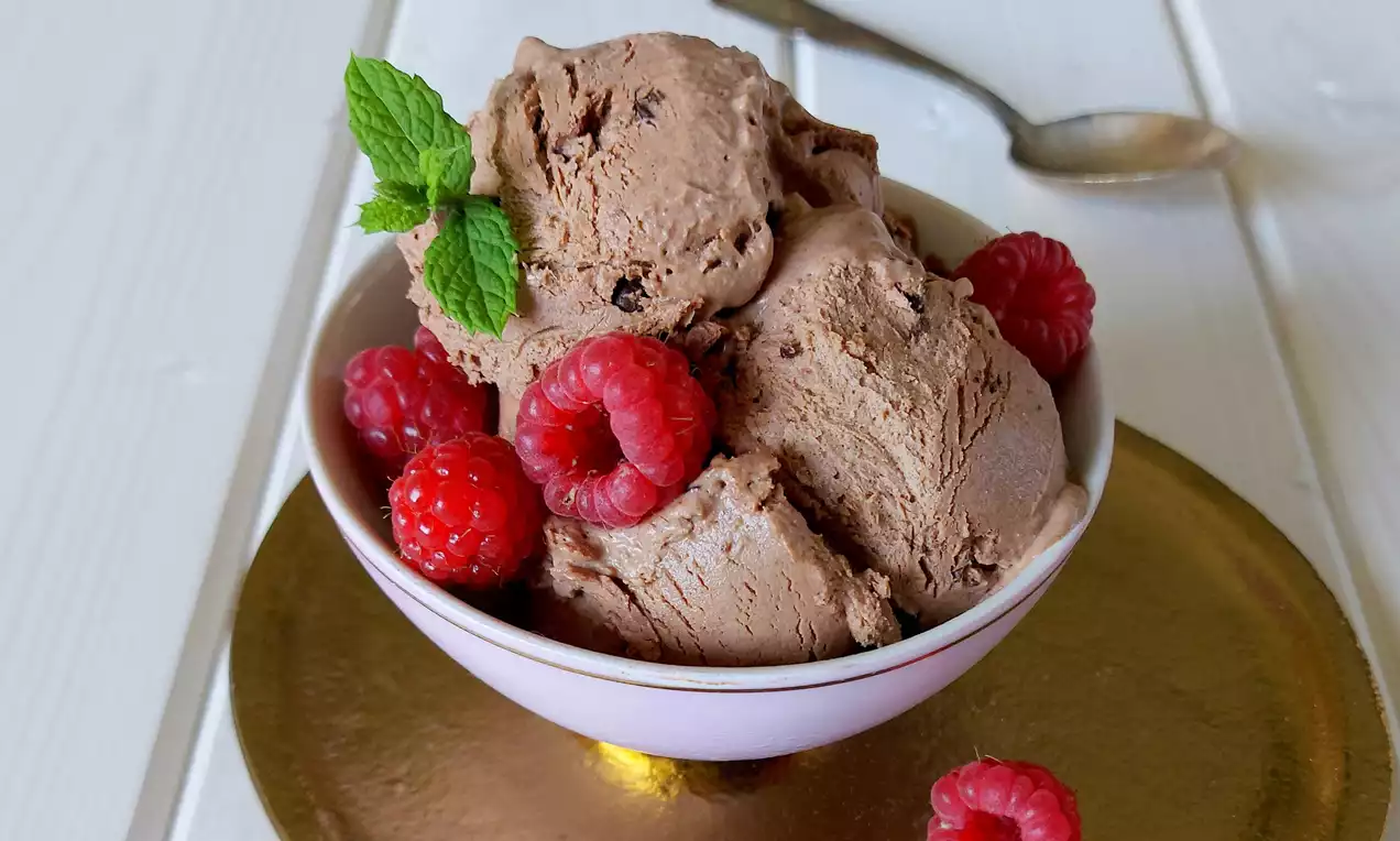 Domácí čokoládová zmrzlina v růžové misce, krémové hnědé barvy posypaná kakaovými boby, ozdobená malinami a lístky máty na zlatém podkladu