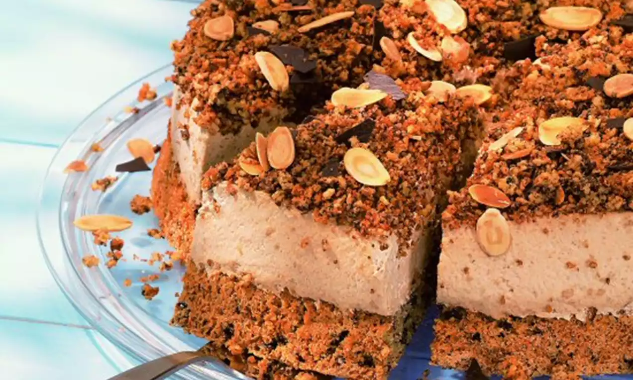 Kávový dort se světlým krémem na tmavém sušenkovém základu, posypaný drobenkou, plátky mandlí a čokoládovými kousky na skleněném servírovacím tácu