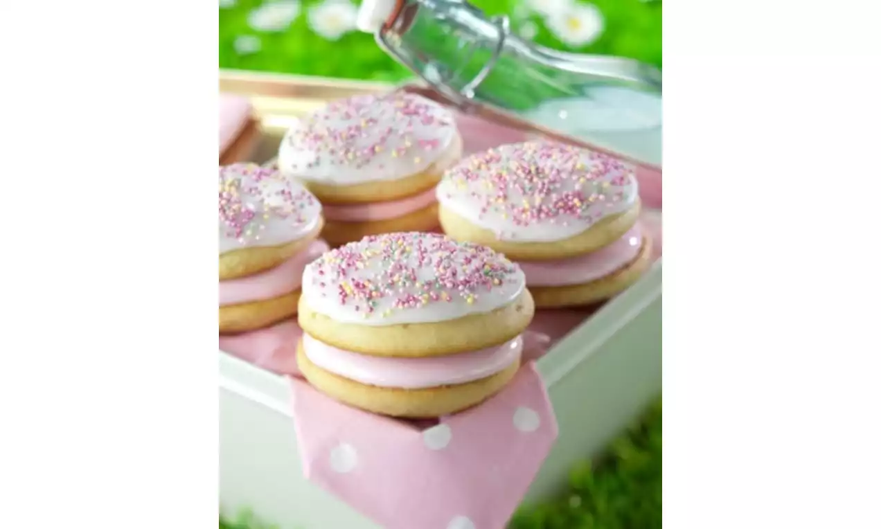 Golden vanilla whoopie pies with pale pink filling, glossy white icing sprinkled with pastel sprinkles, placed on a pink polka dot cloth