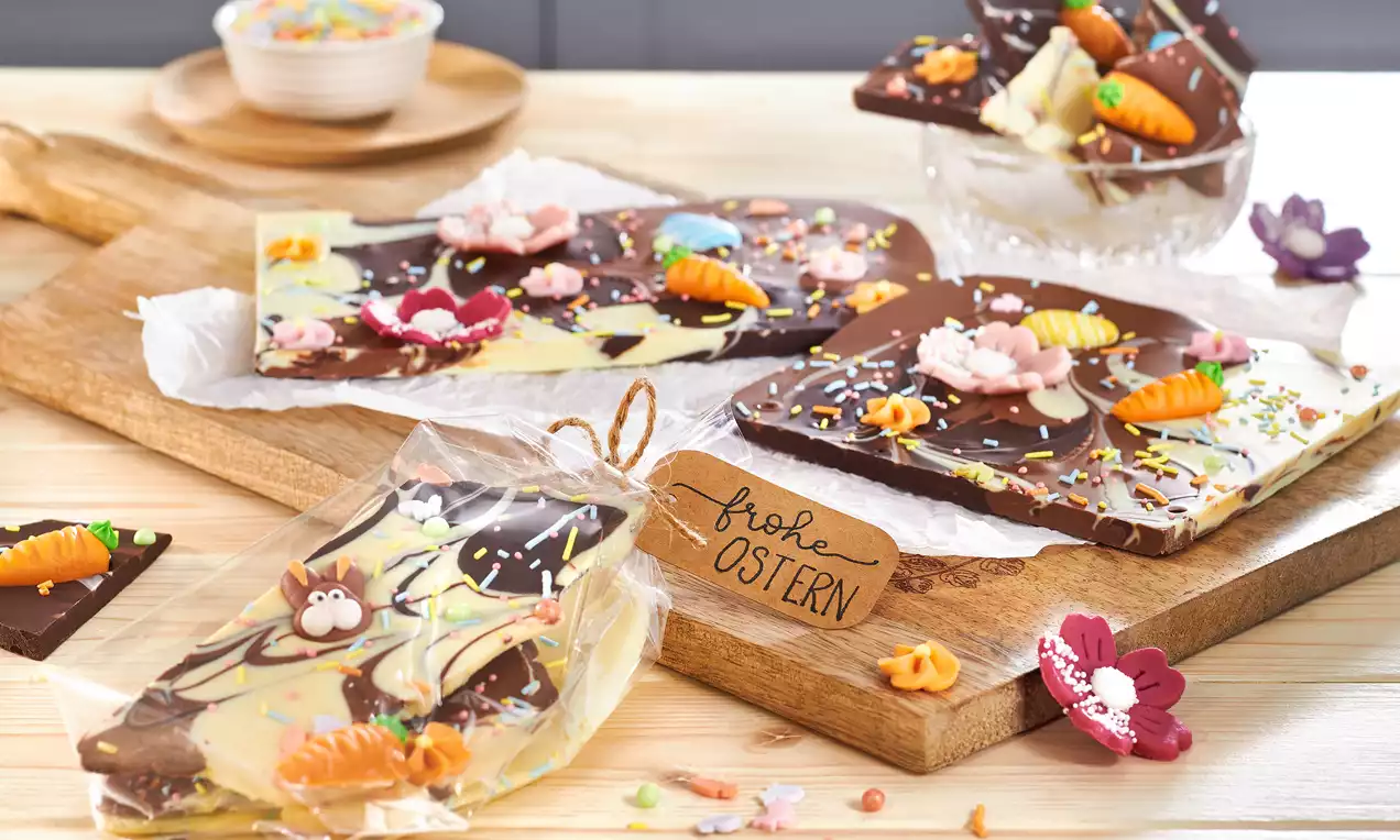 Bunte Bruchschokoladentafeln aus dunkler und heller Schokolade mit Osterdekoration wie Marzipankarotten und Zuckerblüten auf Holzbrett drapiert