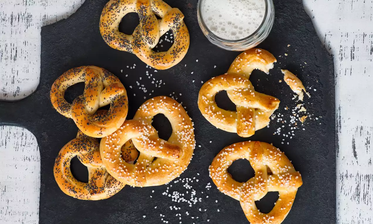 Zlatisté preclíky sypané soľou a makom, usporiadané na tmavej bridlicovej doske vedľa skleného pohára s penivým nápojom