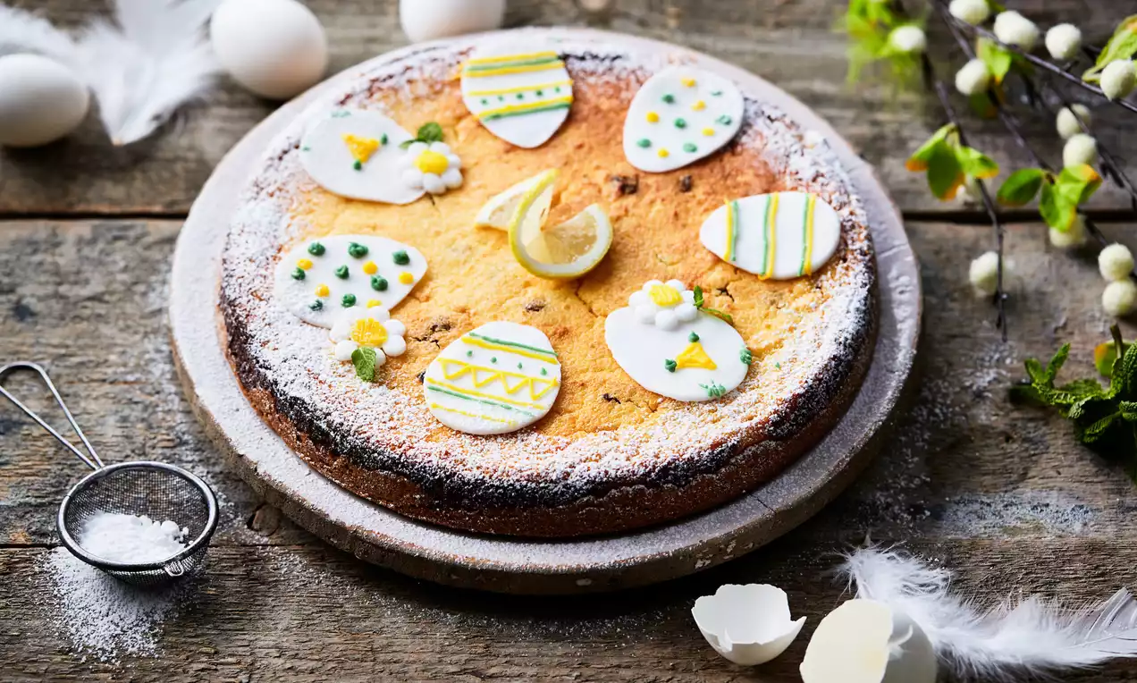 Ronde, goudbruine paastaart met poedersuiker, gedecoreerd met kleurrijke marsepeinen paaseitjes en een schijfje citroen op een rustiek houten oppervlak