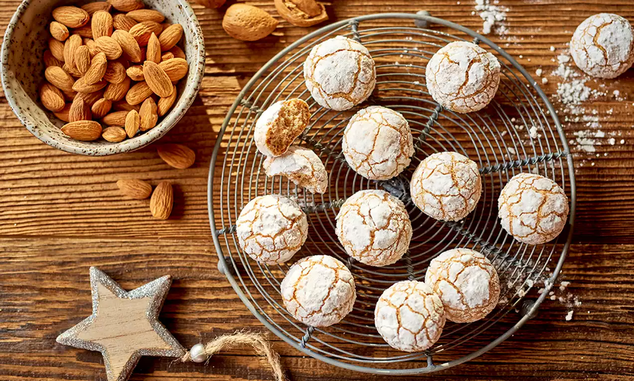 Popękane migdałowe ciasteczka o jasnobrązowej barwie, oprószone cukrem pudrem, ułożone na okrągłej metalowej kratce, obok miski z migdałami