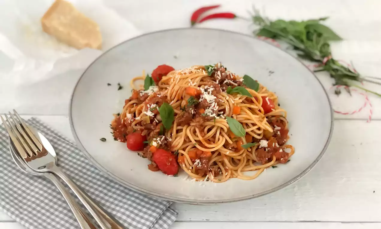 Spaghetti mit rotbrauner Bolognese-Sauce, frischen Kräutern und Parmesan auf einem grauen Teller, dekoriert mit roten Chilischoten und Basilikum