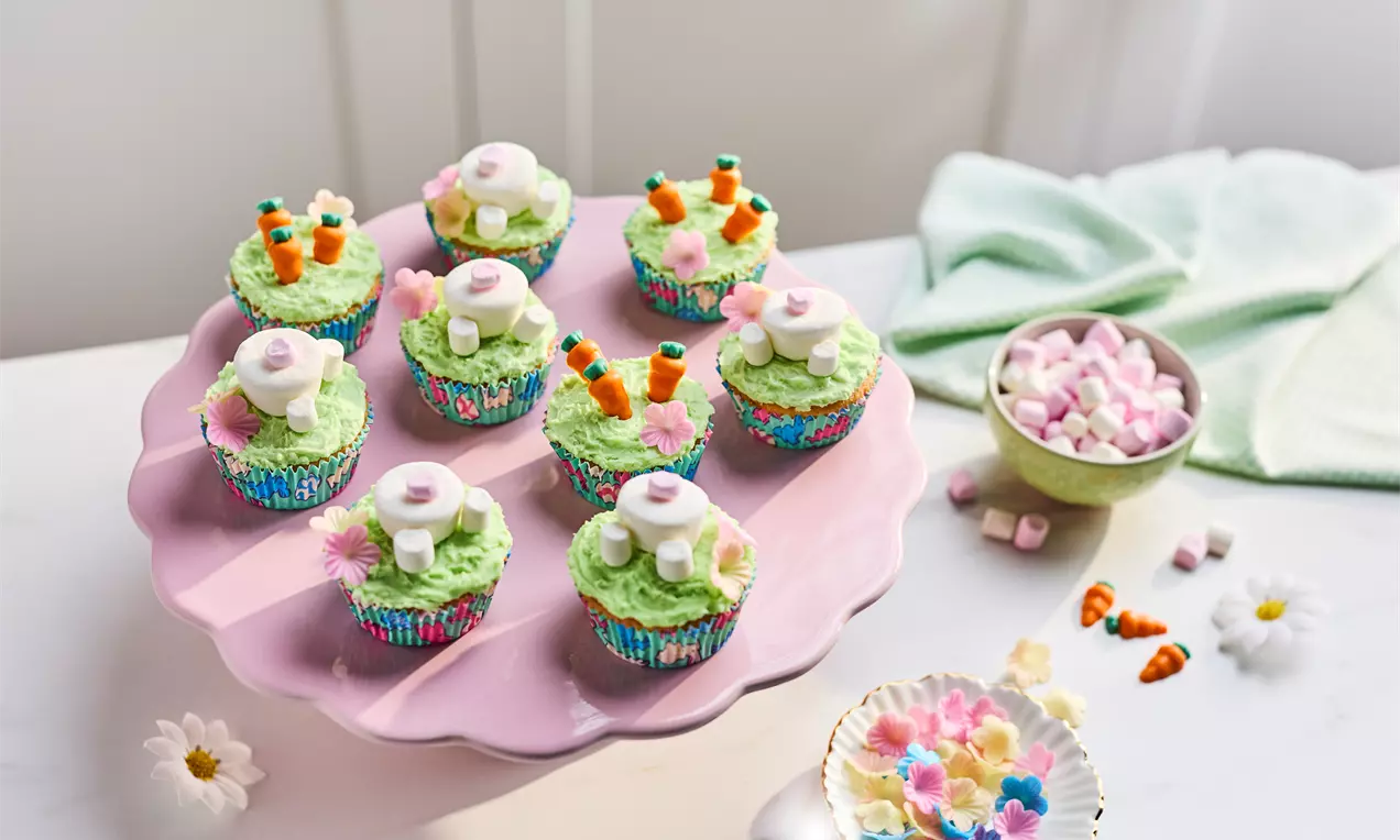 Cupcakes with light green frosting, small bunny-shaped decorations, marshmallow tails, and carrot toppers on a pink stand