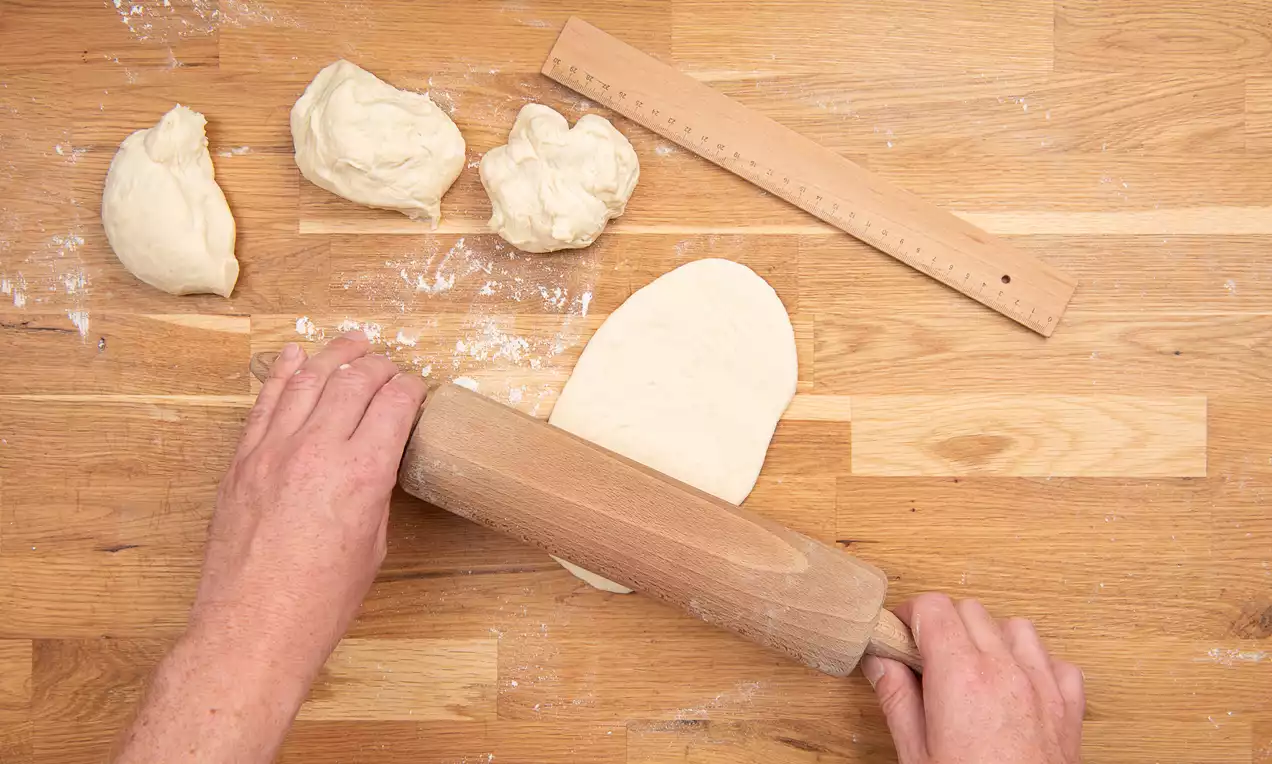 Zwei Hände rollen mit einem hölzernen Nudelholz eine beige Teigkugel auf einer hellen Holzarbeitsfläche zu einem flachen Oval aus