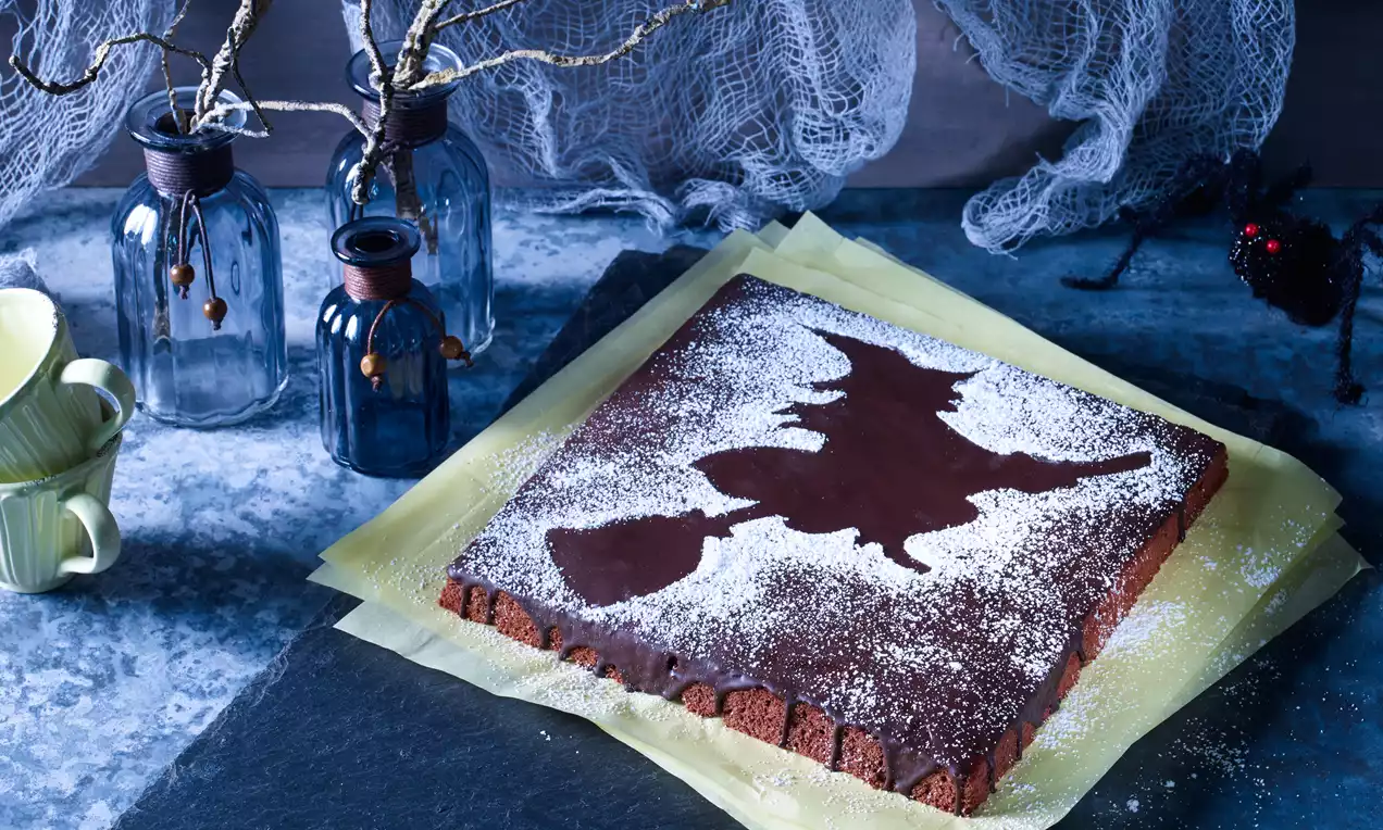 Rechteckiger Schokokuchen mit weißer Puderzucker-Hexensilhouette auf gelben Papierlagen, arrangiert vor Glasflaschen und Spinnweben