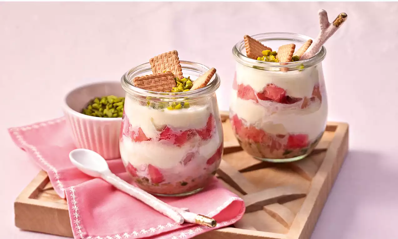 Quark-Frucht-Dessert im Glas mit geschichteter Creme, roten Fruchtstücken, Keksdekor, Pistazien und einer rosa Serviette auf Holztablett