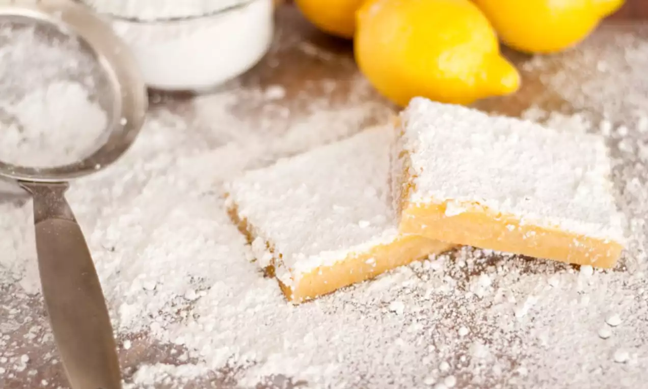 Lemon squares with golden crumbly edges, covered in white powdered sugar, lying on a wooden surface near fresh lemons and a sifter