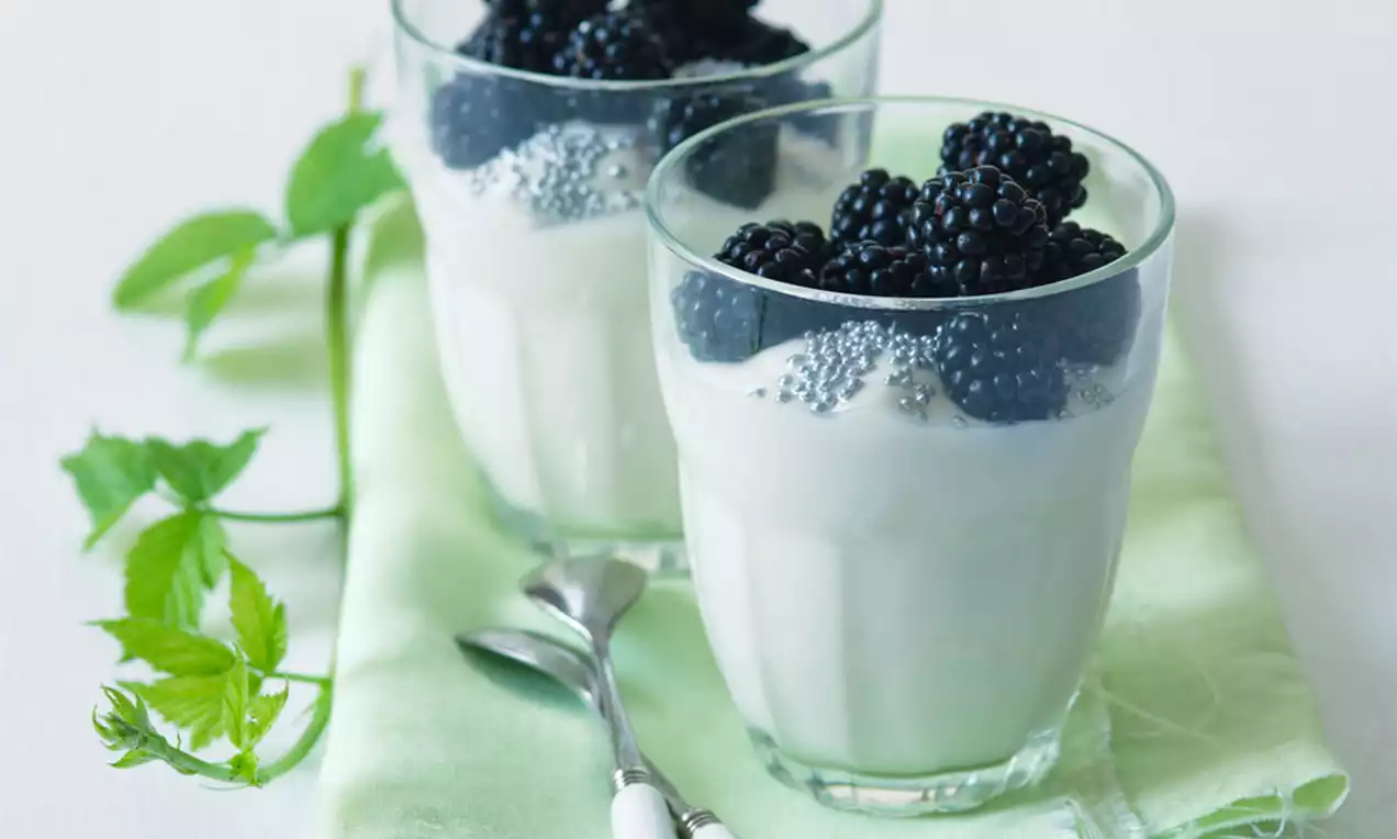 Sklenený pohár so smotanovo bielou náplňou, na ktorej sú usporiadané lesklé čiernofialové ostružiny, položený na svetlozelenej látke