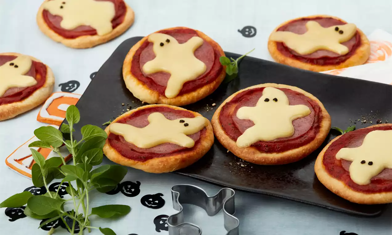 Kleine runde Pizzinis mit rotem Belag und geschmolzenem hellgelbem Käse in Gespensterform auf schwarzem Teller, dekoriert mit Kräutern