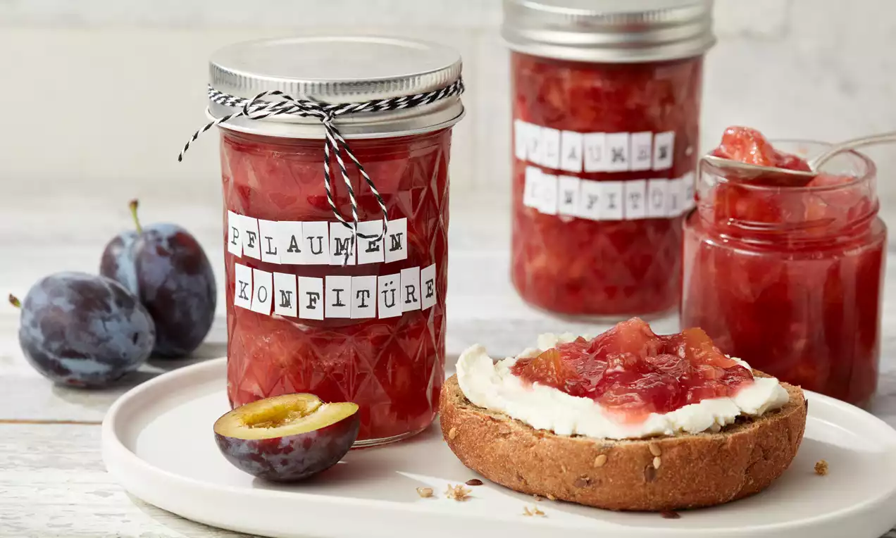 Pflaumenkonfitüre im facettierten Glas mit silbernem Deckel und handgeschriebenen Etiketten, daneben frische Pflaumen und bestrichenes Brötchen