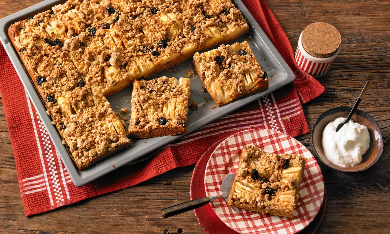 Streusel-Apfel-Birnen-Blechkuchen mit goldbrauner Kruste, saftigen Fruchtstücken und Rosinen auf rotem Karomuster-Teller neben Sahne