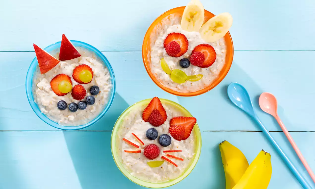 Apfel-Zimt-Porridge in drei bunten Schalen, mit Joghurtcreme, Obstdekor in Tierformen und zwei gelben Bananen auf hellblauem Holztisch