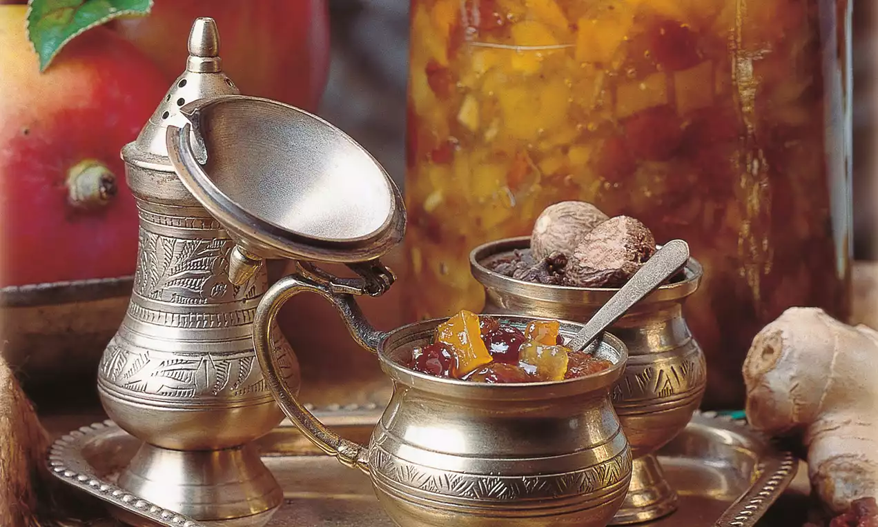 Žiarivo žlté mango-jablkové chutney s lesklou textúrou v ozdobných kovových nádobách na podnose, obklopené detailmi korenia a ovocia