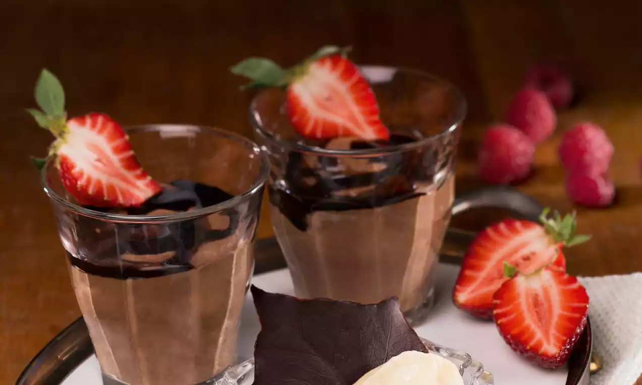 Čokoladni mousse tamnosmeđe boje u prozirnim čašama, ukrašen svježom polovinom jagode i čokoladnim preljevom na bijelom tanjuru