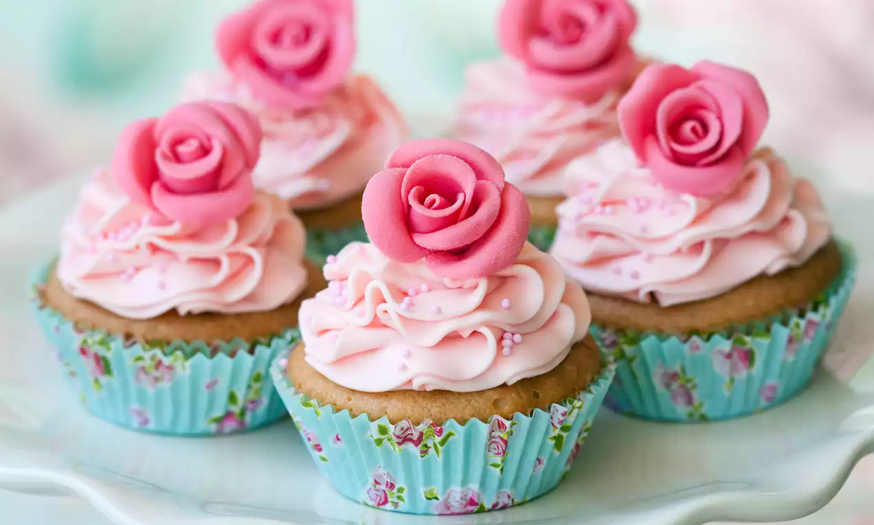 Golden cupcakes in floral wrappers topped with swirls of pale pink frosting, pink sugar pearls, and delicate rose-shaped decorations