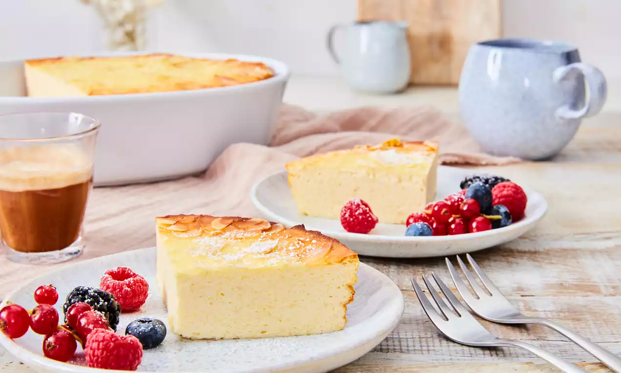 Zwei Stücke goldgelber Quark-Grieß-Auflauf auf weißen Tellern, serviert mit frischen Beeren, daneben Besteck und ein Glas Kaffee