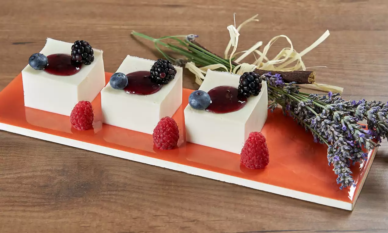 Cubetti bianchi e lisci di panna cotta disposti su un piatto arancione, decorati con frutti di bosco lucidi e rametti di lavanda lilla