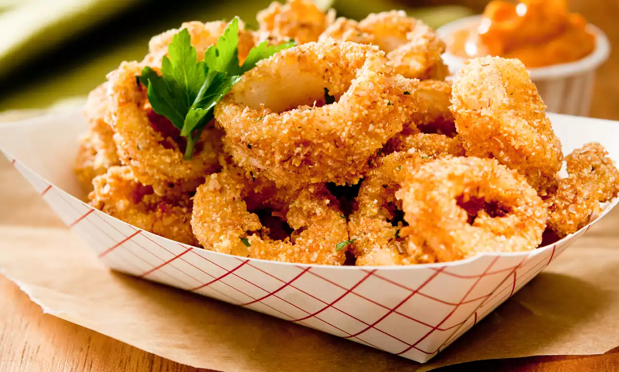 Golden-brown calamari rings with a crunchy oatmeal coating piled in a paper tray, garnished with fresh parsley, sauce in the background