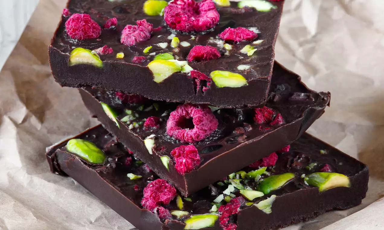 Tmavě hnědé kakaové kvádry s matnou texturou, posypané celými malinami a zelenými pistáciovými kousky, vrstvené na pergamenovém papíře