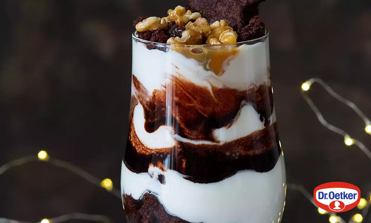 Vysoký pohár s vrstvami tmavohnedej čokoládovej brownie, snehobieleho krému, polevy z karamelu a orechmi na vrchu, na tmavom pozadí