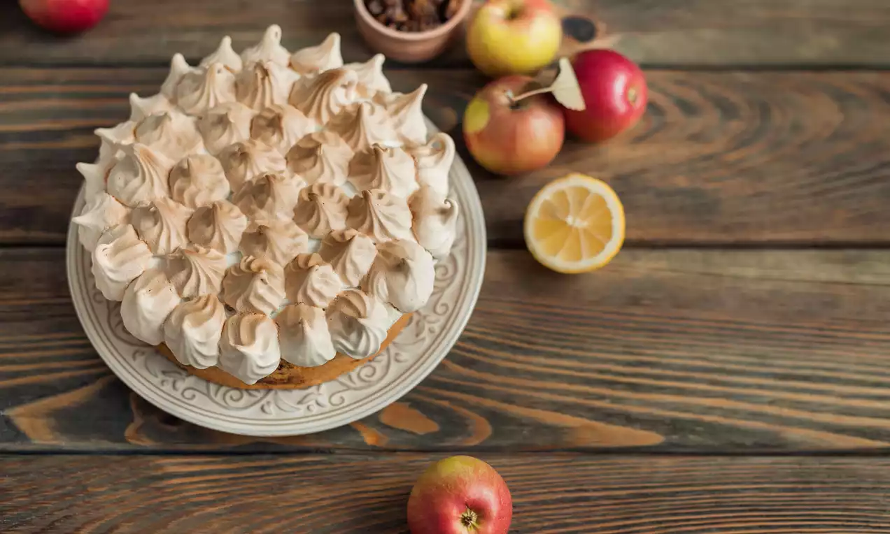 Prăjitură rotundă cu strat auriu de mere moi acoperit cu bezea rumenită în moturi spumoase pe o farfurie decorată, alături de mere și felii de lămâie