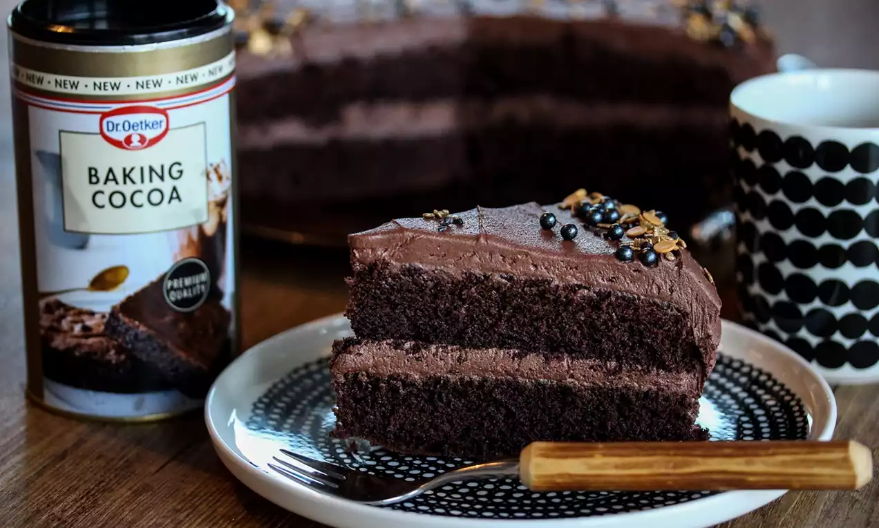 Klassisk sjokoladekake med to mørke lag, sjokoladekrem og perledekor på mønstret tallerken ved siden av kakao og kopp med svart-hvitt mønster