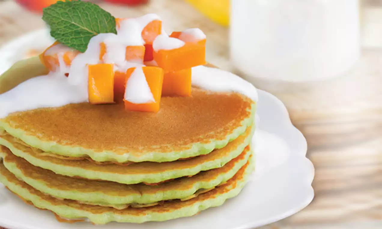 Three golden-brown and green pandan pancakes stacked on a white plate, topped with diced orange mango, white cream, and a green mint leaf