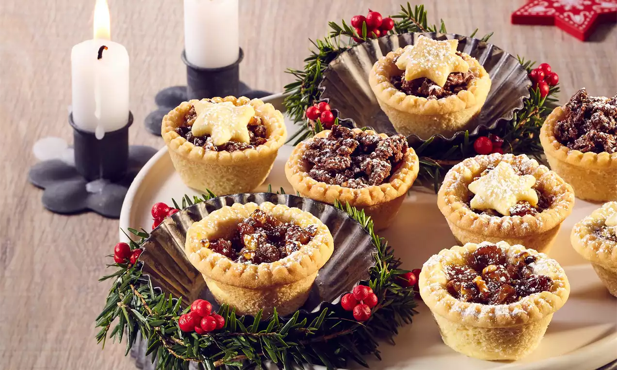 Malé zlatisté košíčky s lesklou ovocnou plnkou, ozdobené hviezdičkami, zasnežené práškovým cukrom, na tanieri s vetvičkami a červenými bobuľami