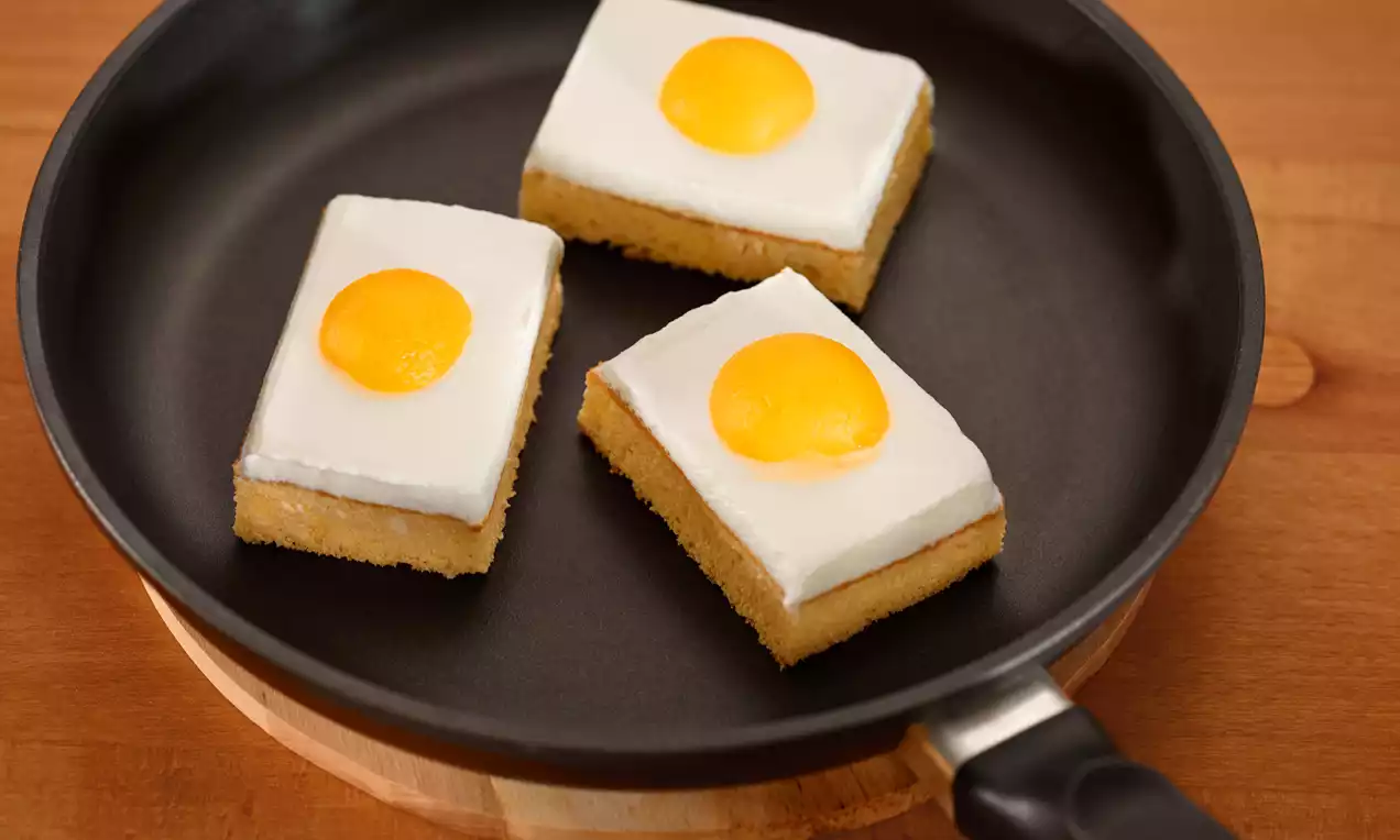 Fette rettangolari di dolce soffice con base dorata, copertura bianca e guarnite con una luminosità arancione simile a tuorli d'uovo in una padella nera