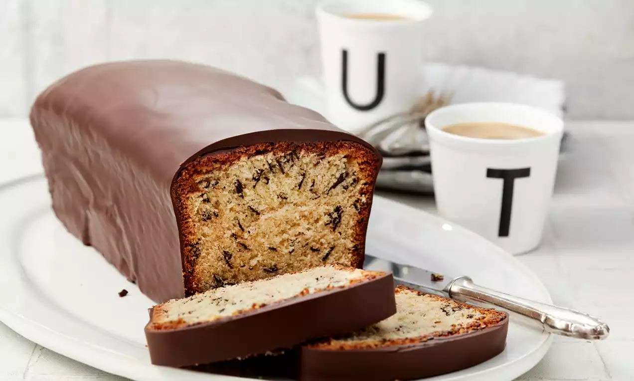 Rechteckiger Marzipankuchen mit dunkler Schokoladen-Glasur, saftigem Teig mit Schokostückchen, auf weißer Platte mit eingeknicktem Messer